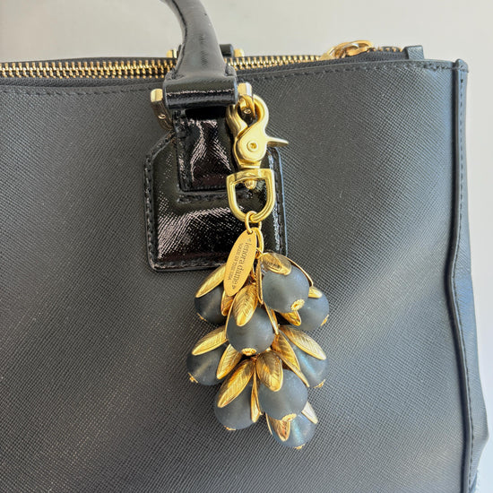 Beaded bag charm featuring matte black glass beads encased in gold plated brass floral leaf shaped bead caps. Gold trigger clasp and an oval hang tag that says Lenora Dame Made in USA. Lenora Dame bag charm shown on the strap of a black leather handbag.