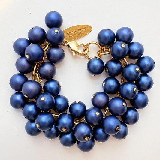 A gold chain bracelet with multiple strands of matte navy blue faux pearls. The bracelet has a small tag that reads 'Lenora Dame' and 'Made in the USA.'