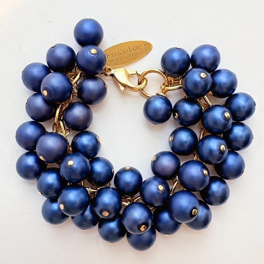 A gold chain bracelet with multiple strands of matte navy blue faux pearls. The bracelet has a small tag that reads &#39;Lenora Dame&#39; and &#39;Made in the USA.&#39;