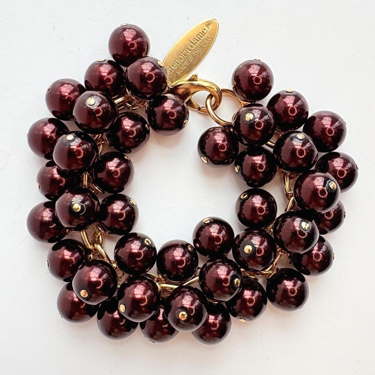 A gold chain bracelet with multiple strands of shiny dark burgundy faux pearls. The bracelet has a small tag that reads 'Lenora Dame' and 'Made in the USA.'