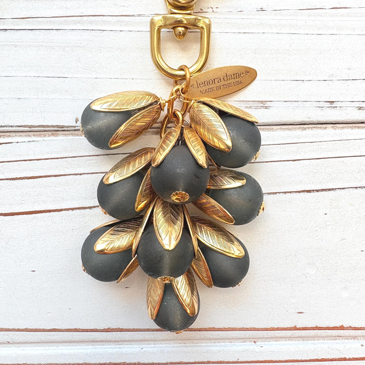 Beaded bag charm featuring matte black glass beads encased in gold plated brass floral leaf shaped bead caps. Gold trigger clasp and an oval hang tag that says Lenora Dame Made in USA. Lenora Dame bag charm.
