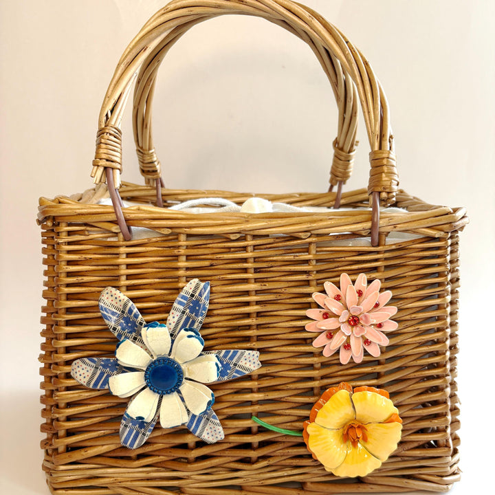 Top handle rectangular woven wicker handbag embellished with three metal flower brooches. One flower is blue and white, one pink flower brooch and one yellow and orange flower brooch. White fabric inner lining. Cute handbags for summer.