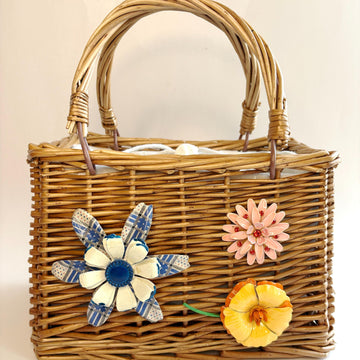Top handle rectangular woven wicker handbag embellished with three metal flower brooches. One flower is blue and white, one pink flower brooch and one yellow and orange flower brooch. White fabric inner lining. Cute handbags for summer.