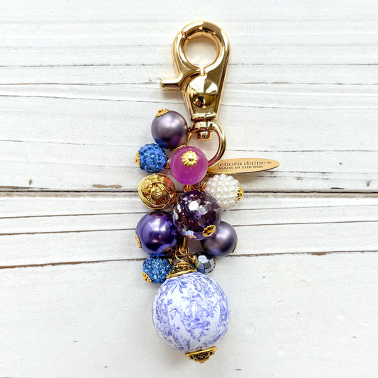 Beaded bag charm with a mix of bead styles in shades of purple, blue, gold and pearl. One larger round bead in a purple and white toile print hangs at the bottom and a gold trigger clasp on top of all of the beads. Brass oval hang tag near the trigger clasp that says "Lenora Dame Handmade in USA". Lenora Dame bag charm.