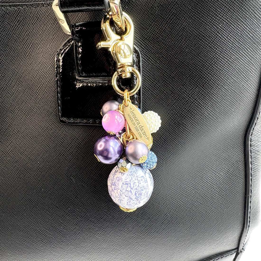Beaded bag charm with a mix of bead styles in shades of purple, blue, gold and pearl. One larger round bead in a purple and white toile print hangs at the bottom and a gold trigger clasp on top of all of the beads. Brass oval hang tag near the trigger clasp that says &quot;Lenora Dame Handmade in USA&quot;. Lenora Dame bag charm.