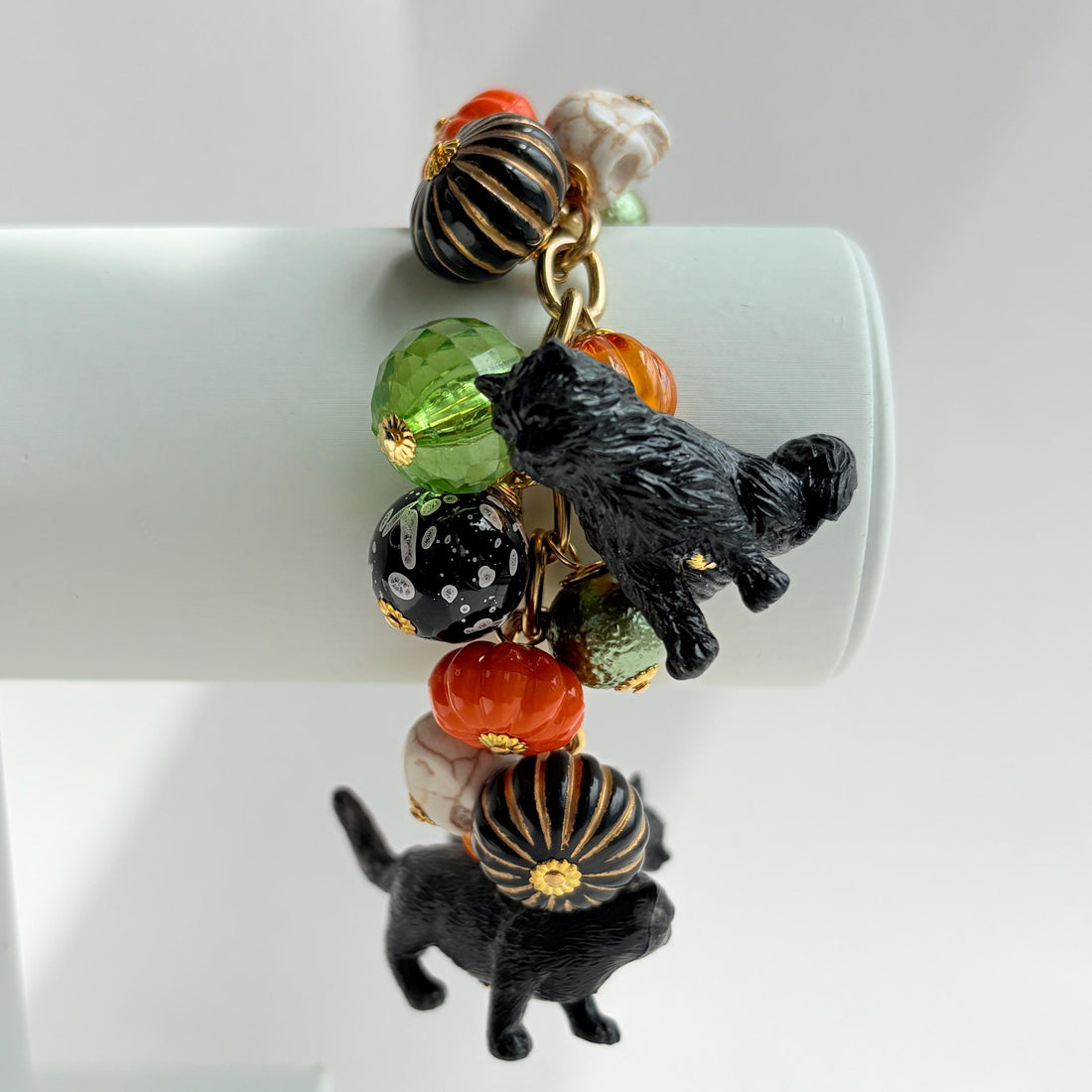 A gold chain bracelet with black cat charms, green and orange beads, and a white pumpkin charm. The bracelet has a tag that reads &