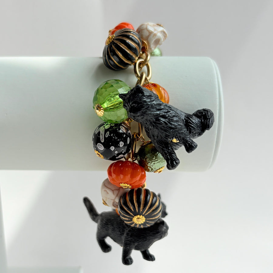 A gold chain bracelet with black cat charms, green and orange beads, and a white pumpkin charm. The bracelet has a tag that reads &#39;Lenora Dame&#39; and &#39;Made in USA&#39;.