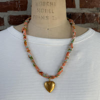 May include: A necklace with a peach-colored fabric cord, adorned with floral patterns in green, blue, and yellow. A large, gold-colored heart pendant hangs from the center, creating a vintage-inspired accessory.