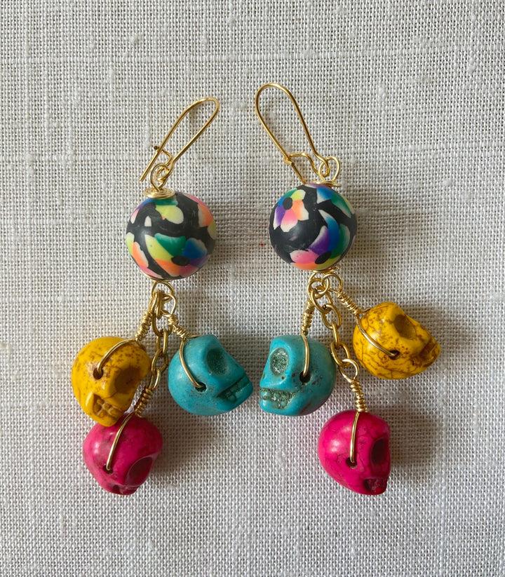 Lenora Dame sugar skull earrings with three miniature carved skulls in bright yellow, hot pink and turquoise wire wrapped under a black with rainbow flower design round polymer bead dangling from a gold French ear wire.