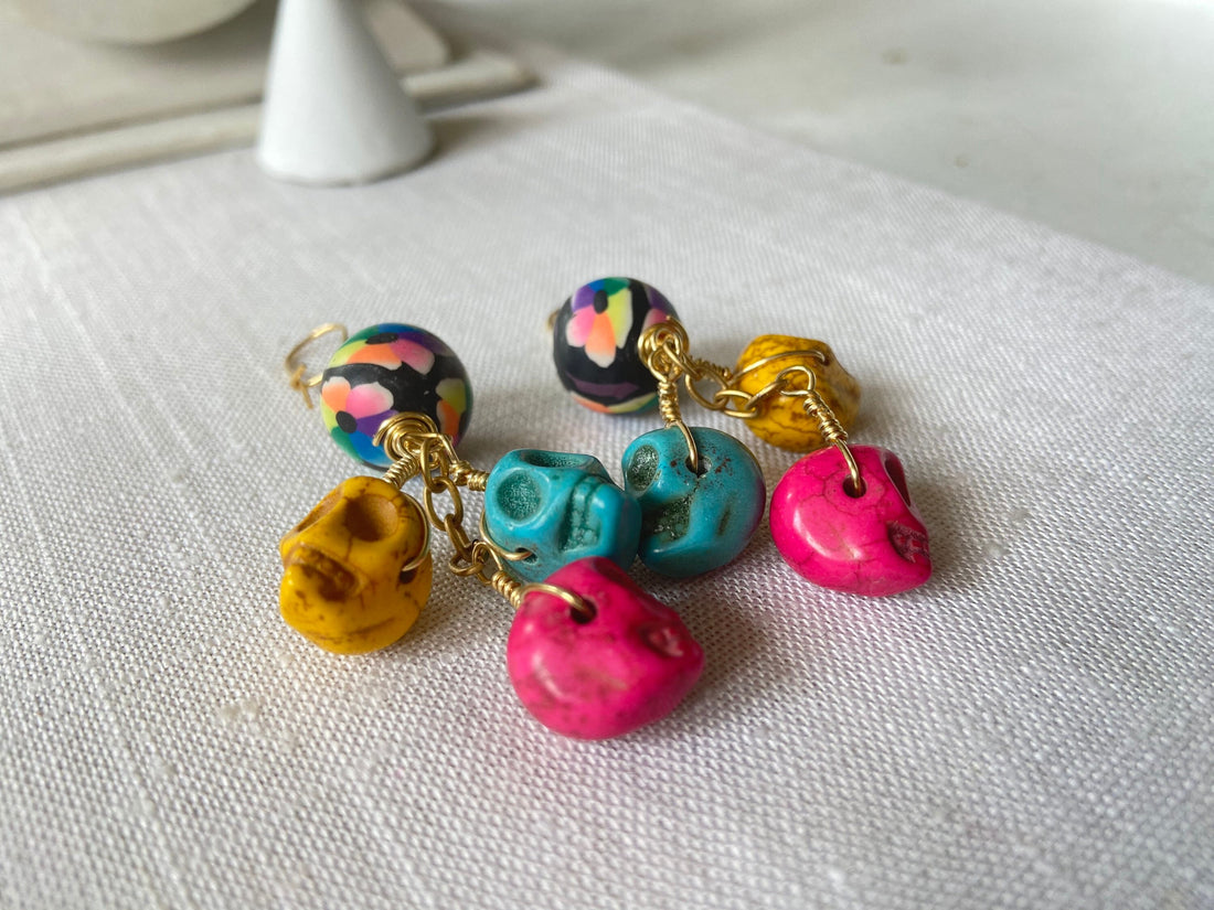 Lenora Dame sugar skull earrings with three miniature carved skulls in bright yellow, hot pink and turquoise wire wrapped under a black with rainbow flower design round polymer bead dangling from a gold French ear wire. 