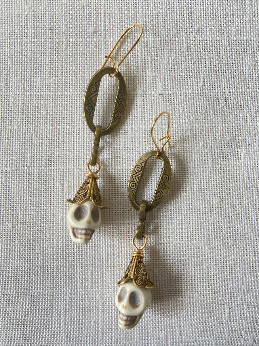 Lenora Dame Shackled Skull Earrings with white carved sugar skulls topped with brass filigree cone bead dangling from oval etched metal pieces in an antique bronze finish. Earrings hang from gold French hook ear wires. 