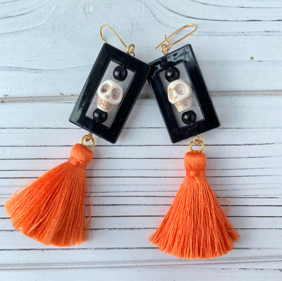 Lenora Dame Halloween earrings with white carved sugar skulls framed in black shiny rectangular frames stacked above bright orange silky tassels. Earrings are hung on gold French hook ear wires.