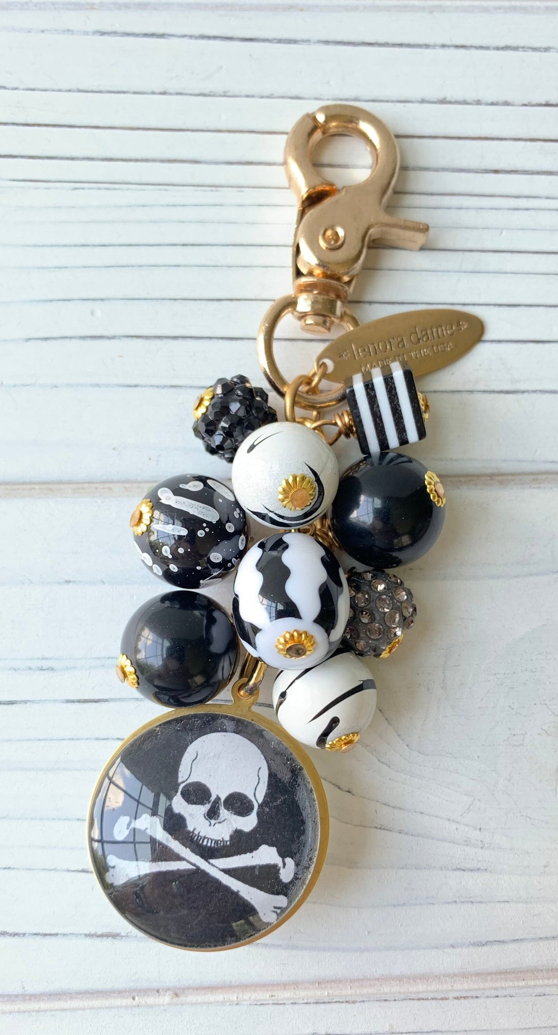 Lenora Dame Halloween purse charm with a mix of different patterned black and white beads wire wrapped on a gold-plated cable chain. A white skull and crossbones with black background picture under a domed glass lens mounted on a round brass charm hangs from the bottom. Gold trigger clasp is at the top.