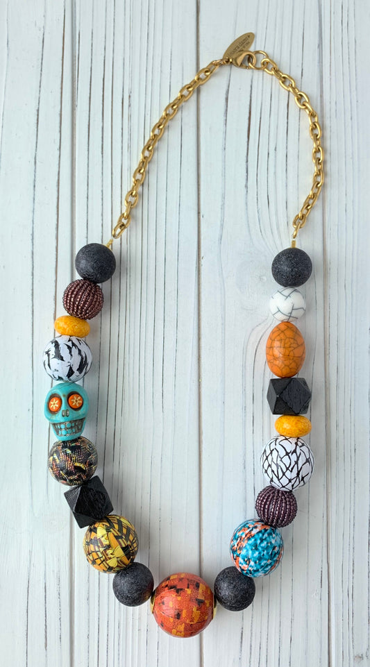 Lenora Dame Halloween necklace with a mix of oversized beads strung together and attached to gold-plated cable chain with lobster clasp. The large round beads include black foil texture, purple seed bead balls, yellow spacers, black squares and wooden balls decoupaged n a mix of vintage textile prints. A turquoise colored carved sugar skull with orange and gold star eyes is strung among the beads on the left side.