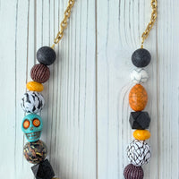 Lenora Dame Halloween necklace with a mix of oversized beads strung together and attached to gold-plated cable chain with lobster clasp. The large round beads include black foil texture, purple seed bead balls, yellow spacers, black squares and wooden balls decoupaged n a mix of vintage textile prints. A turquoise colored carved sugar skull with orange and gold star eyes is strung among the beads on the left side.  