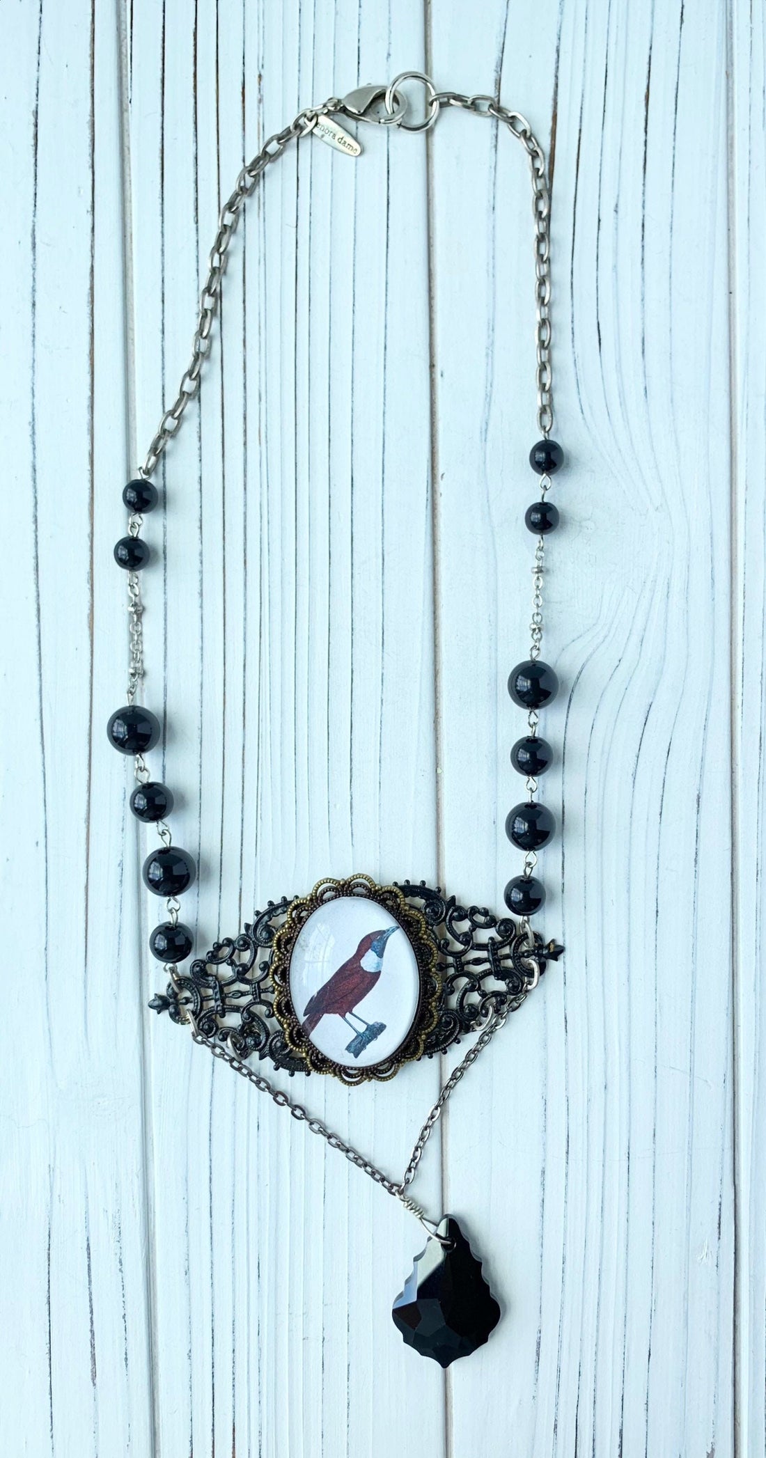 Lenora Dame necklace with a raven picture under a domed glass lens mounted on two layers of brass filigree hand painted black that hangs between silver chain with round shiny black beads. A black faceted irregular shaped teardrop glass pendant hangs from two layers of silver chain under the raven pendant.