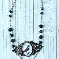 Lenora Dame necklace with a raven picture under a domed glass lens mounted on two layers of brass filigree hand painted black that hangs between silver chain with round shiny black beads. A black faceted irregular shaped teardrop glass pendant hangs from two layers of silver chain under the raven pendant.