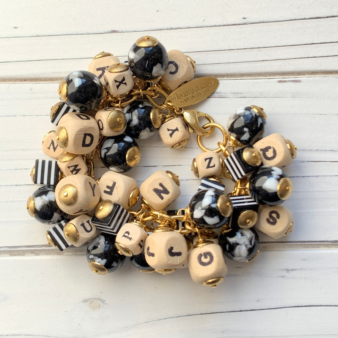 Chunky charm bracelet filled with wooden letter beads, striped square beads and round black and white beads hung on a gold cable chain with lobster clasp and Lenora Dame hang tag. 