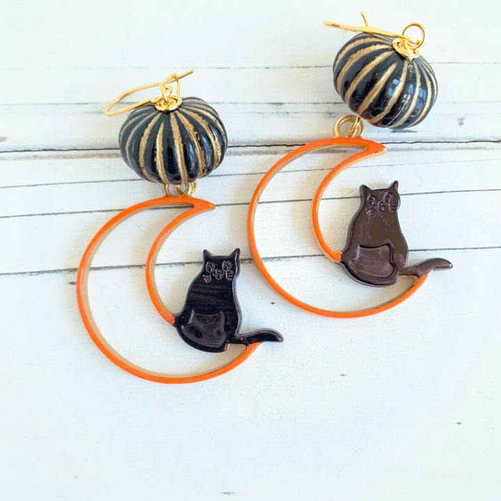 Lenora Dame Halloween earrings with hand-painted orange and gold metal crescent moon cutout with black cat sitting on the edge. The crescent moons hang from miniature black with gold stripes pumpkins. 