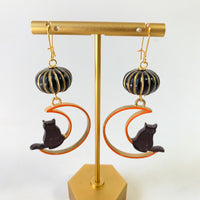 Lenora Dame Halloween earrings with hand-painted orange and gold metal crescent moon cutout with black cat sitting on the edge. The crescent moons hang from miniature black with gold stripes pumpkins.