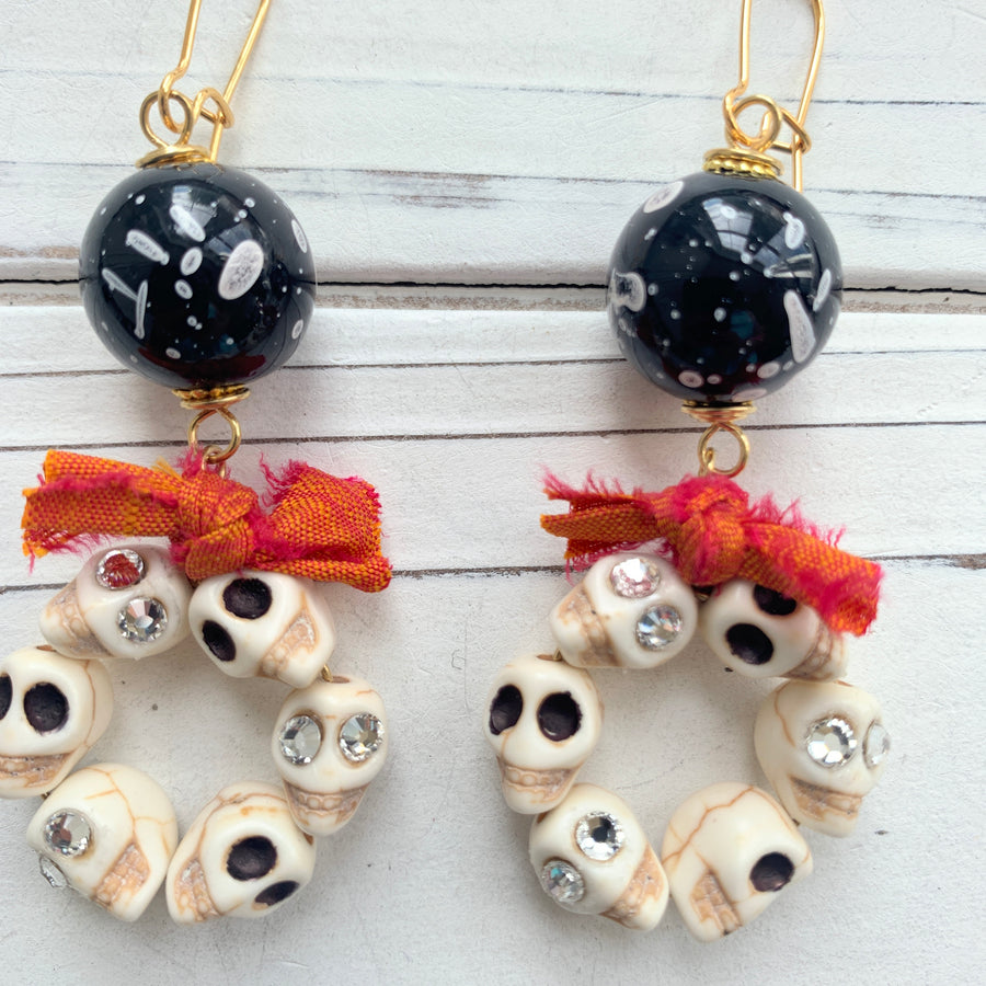 Lenora Dame Halloween earrings with white miniature carved sugar skulls, some with rhinestone embellished eyes, shaped into hoops and adorned with a bow made from orange and red sari silk. The skull hoops dangle from black with white splatter acrylic beads attached to gold-plated French hook ear wires.